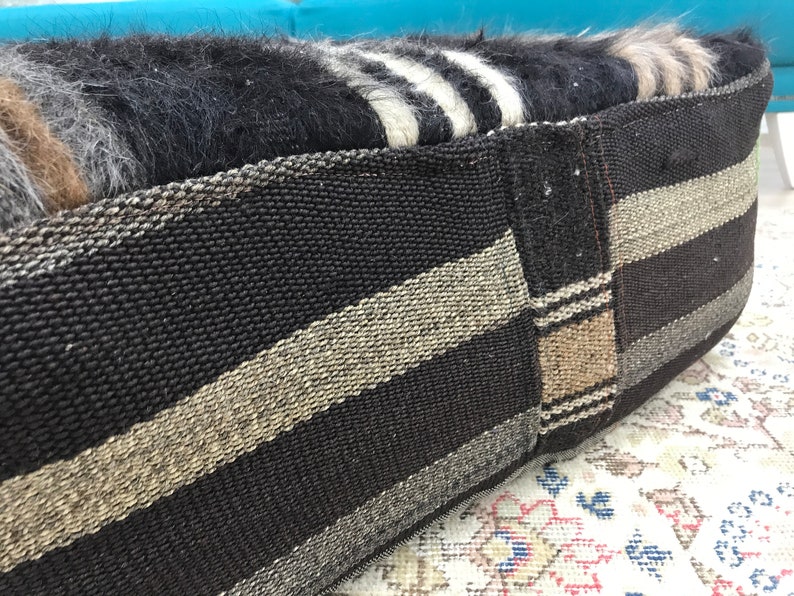 May include: A black and white striped woven cushion with a brown and beige striped panel. The cushion is made of a soft, fuzzy material.