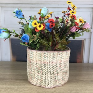 May include: A woven basket with a pink and green pattern holds a bouquet of artificial flowers. The flowers are blue, yellow, red, and pink.