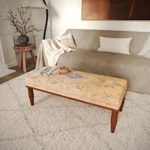 May include: A rectangular ottoman with a floral patterned top in shades of yellow, blue, and green. The ottoman has a dark brown wooden frame and legs. It is placed on a white shag rug in a living room setting.