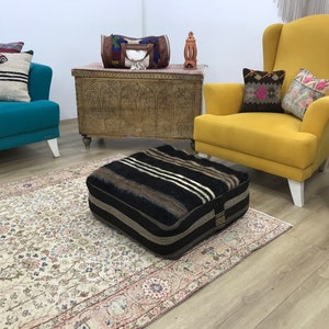 May include: A black and white striped floor cushion with a brown fringe sits on a cream colored rug in a living room setting. A yellow armchair with white legs and a teal sofa with white legs are in the background.