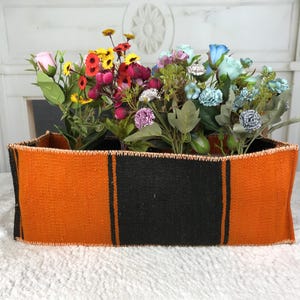 Puede incluir: Una cesta rectangular tejida con un vibrante patrón de rayas naranjas y negras con costuras visibles a lo largo de los bordes superior e inferior, llena de un surtido de flores artificiales en tonos rosa, amarillo, rojo, morado y azul.