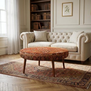 May include: A round ottoman featuring a detailed, multi-colored oriental rug pattern in shades of deep red, orange, and navy blue. The ottoman is supported by four tapered wooden legs and is positioned in front of a cream-colored tufted sofa.