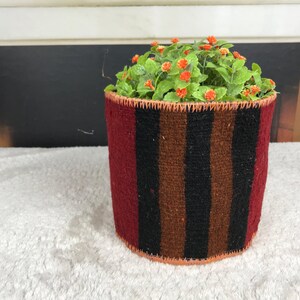 May include: A cylindrical woven fabric planter cover featuring vertical stripes in deep red, dark brown, and black. The top and bottom edges are finished with a visible orange whipstitch detail. A small green plant with tiny orange flowers sits inside.