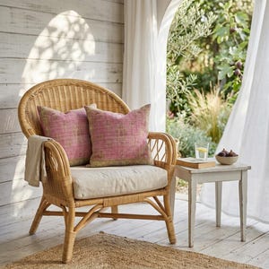 May include: A wicker chair with two pink throw pillows with gold trim. A beige throw blanket rests on the chair. A small white side table holds a glass of water, a book, and a bowl of figs. The scene is set on a porch.
