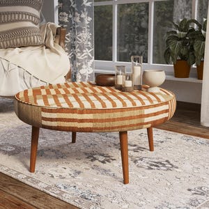 May include: A round coffee table featuring a textured, woven fabric top with a geometric pattern in shades of rust orange, cream, and tan. The table is supported by four tapered, medium-brown wooden legs and has a low-profile, cylindrical shape.