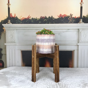 May include: A knitted planter pot with a striped pattern in shades of lavender, beige, and white, filled with greenery and red berries. It sits on a dark brown wooden plant stand. The background features a fireplace with a Christmas garland.