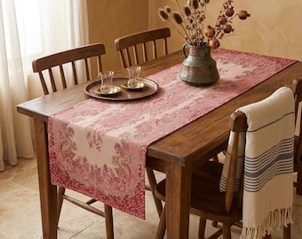 Op de afbeelding: Een houten eettafel met een roze bloemen tafelloper. Twee heldere glazen en een kleine dienblad staan op de tafel. Een vaas met gedroogde bloemen en een opgevouwen handdoek op een stoel maken de scène compleet.