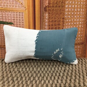 May include: Rectangular decorative pillow with a two-tone design. One half is white, the other is teal. The teal section features white stitching and a decorative stitched design. The pillow is placed on a textured, woven surface.