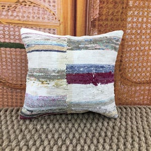 Pode incluir: Almofada decorativa quadrada com um design de patchwork. A almofada apresenta uma mistura de tecido branco, azul, bordô e tons neutros. A almofada é colocada sobre uma superfície tecida texturizada.