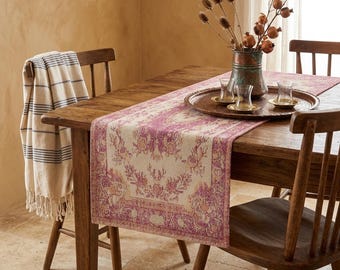 Op de afbeelding: Een houten eettafel met een bloemenpatroon tafelloper in roze en beige. Een koperen dienblad met glazen en een vaas met gedroogde bloemen staat op de tafel. Een gestreepte deken is over een houten stoel gedrapeerd.