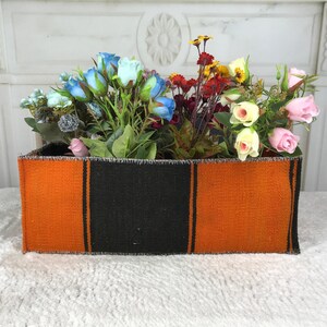 May include: A rectangular fabric storage bin featuring a bold striped pattern in alternating blocks of burnt orange and black. The container is filled with an assortment of artificial flowers in shades of blue, red, yellow, cream, and soft pink.