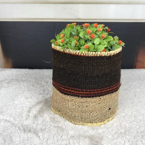 May include: A cylindrical woven basket planter featuring a dark brown upper section and a natural tan lower section, separated by a thin red and black striped band. The basket contains a cluster of green foliage with small orange flowers.