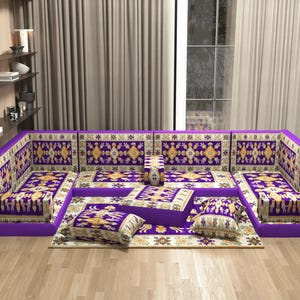 May include: A large, modular purple sofa with patterned cushions and a matching rug. The sofa features a traditional design with gold and white accents. The setup includes multiple cushions and a central ottoman, creating a comfortable seating area.