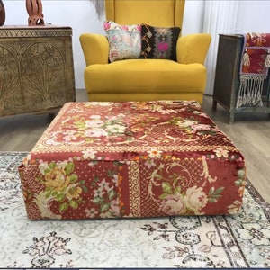 Pode incluir: Um almofada de ch&atilde;o de veludo vermelho com um padr&atilde;o floral. O almofada &eacute; quadrado e tem um topo acolchoado. Est&aacute; sentado em um tapete em uma sala de estar.