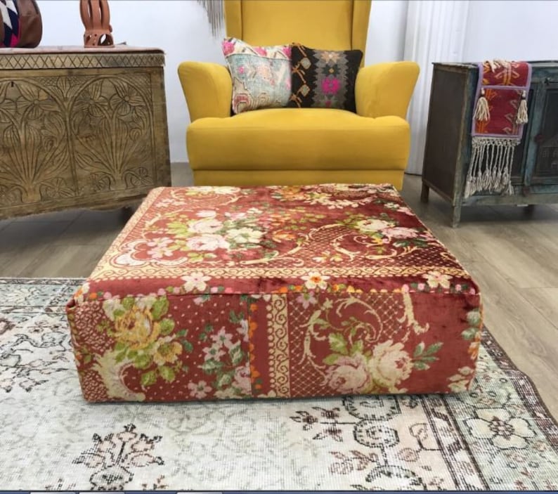Pode incluir: Um almofada de ch&atilde;o de veludo vermelho com um padr&atilde;o floral. O almofada &eacute; quadrado e tem um topo acolchoado. Est&aacute; sentado em um tapete em uma sala de estar.