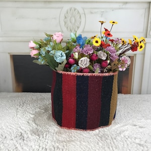 May include: A woven basket with red, black, and brown stripes holds a bouquet of pink, purple, yellow, and blue artificial flowers.