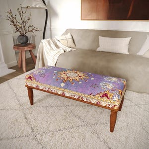 May include: A rectangular ottoman featuring a purple fabric top with a central yellow and white floral medallion pattern, accented by a decorative border in shades of red, gold, and blue, supported by four turned wooden legs.