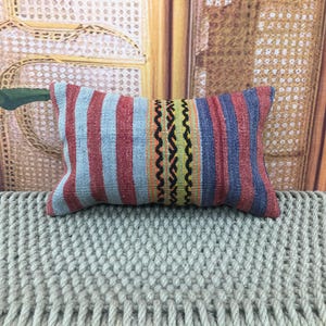 May include: Rectangular decorative pillow with a striped pattern in red, blue, and light blue. The center features a design with black, yellow, and orange zigzags. The pillow is placed on a textured, light gray surface.