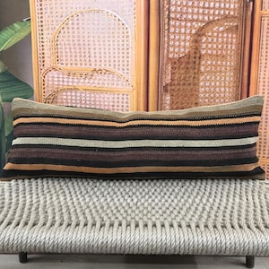 Puede incluir: Un cojín rectangular con rayas horizontales en tonos marrón, negro y beige. El cojín está colocado sobre un banco tejido con un diseño de cuerda clara. El fondo presenta una pantalla de madera y hojas verdes.