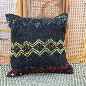 Puede incluir: Un cojín decorativo cuadrado negro con un patrón geométrico en amarillo, rojo y turquesa. El cojín tiene un aspecto texturizado y probablemente está hecho de tela tejida. El diseño presenta formas de zigzag y diamante.