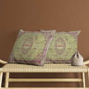 以下が含まれることがあります： 緑と紫の花柄の正方形の装飾枕が2つ、織りのベンチに置かれています。小さな巾着袋が枕の右側にあります。枕はヴィンテージで装飾的なデザインです。