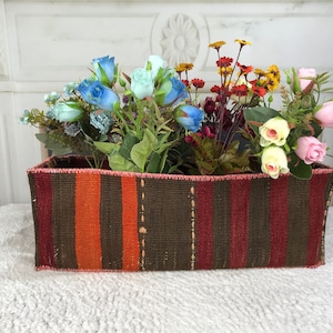 May include: A woven basket with orange, brown and red stripes holds a variety of artificial flowers, including blue roses, pink roses and yellow flowers.