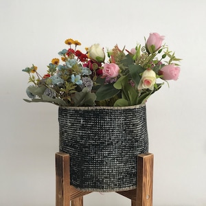 May include: A cylindrical black and white woven fabric basket sits atop a four-legged wooden stand. The basket is filled with an arrangement of artificial flowers, including pink roses, blue blossoms, and small yellow and red wildflowers.
