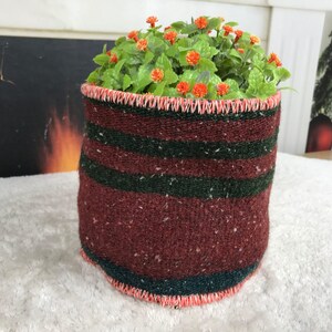 Op de afbeelding: Een cilindervormige stoffen plantenpotcover met horizontale strepen in donkerbordeaux en bosgroen. De boven- en onderranden zijn afgewerkt met een fel koraalkleurige festonsteek. Een kleine groene plant met piepkleine oranje bloemetjes staat erin.