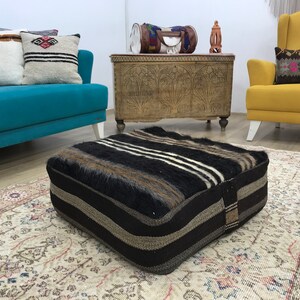 May include: A black and white striped floor cushion with brown accents. The cushion is made of a soft, fuzzy material and is sitting on a patterned rug in a living room setting.