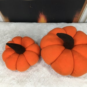 May include: Two orange plush pumpkins with black stems. The larger pumpkin is about 10 inches in diameter and the smaller pumpkin is about 6 inches in diameter.