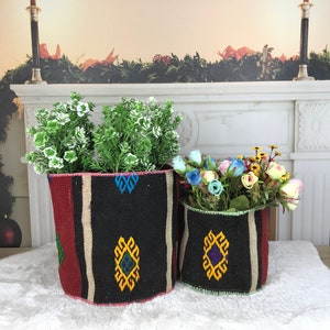 Op de afbeelding: Twee geweven manden met geometrische patronen in rood, zwart, wit en geel. De manden zijn gevuld met groene en blauwe kunstplanten en bloemen.