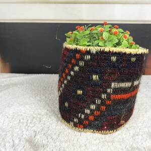 May include: A woven basket planter with a geometric pattern in shades of brown, blue, orange, and white. The planter is filled with a small green plant with red flowers.