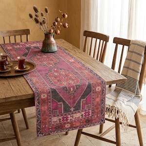 Puede incluir: Una mesa de comedor de madera con un camino de mesa estampado en tonos rosa, rojo y negro. La mesa está decorada con una bandeja con vasos de té y un jarrón con flores secas. Una manta a rayas está sobre una silla. El camino de mesa tiene un diseño de alfombra tradicional.