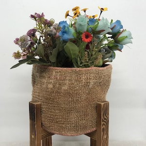 May include: A textured tan woven basket sits inside a dark brown wooden stand. The basket is filled with an arrangement of artificial flowers in shades of blue, purple, yellow, and red, featuring green leaves and stems throughout the bouquet.