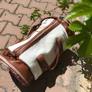 Puede incluir: Una bolsa de lona blanca y marr&oacute;n con cierre de cremallera y asas de cuero. La bolsa est&aacute; tumbada en un camino de adoquines.