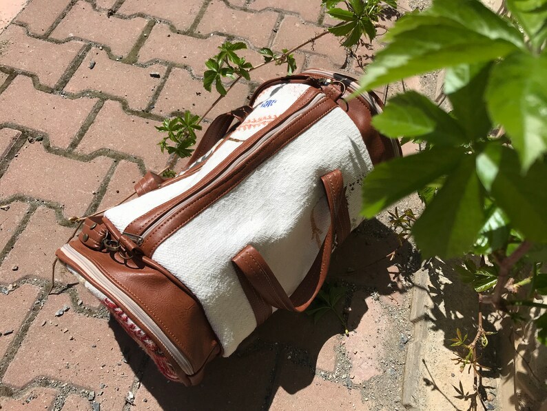 Puede incluir: Una bolsa de lona blanca y marr&oacute;n con cierre de cremallera y asas de cuero. La bolsa est&aacute; tumbada en un camino de adoquines.