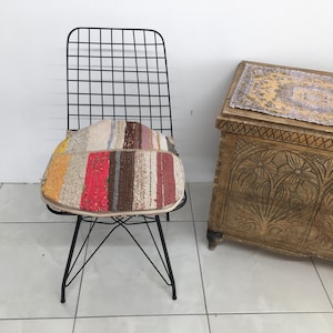 Può includere: Una sedia in metallo nero con schienale a griglia e un cuscino patchwork colorato. Il cuscino presenta strisce verticali in vari colori, tra cui giallo, rosso e marrone. Sullo sfondo, un baule in legno con motivi floreali intagliati.