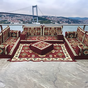 Op de afbeelding: Een traditionele Turkse zithoek met een bordeauxrode en crèmekleurige stof met patronen. De set bevat een grote bank, twee kleinere banken, vier kussens, een salontafel en een tapijt. De set is op een balkon geplaatst met uitzicht op de skyline van de stad.