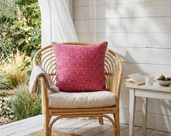 Puede incluir: Una silla de ratán con un cojín cuadrado rosa estampado y una almohada de lino beige. Una manta beige está drapeada sobre el brazo. La silla se encuentra sobre una alfombra tejida. Una pequeña mesa blanca contiene un vaso de agua, un cuenco de higos y un libro abierto.