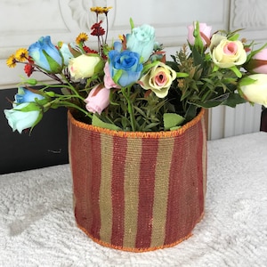 May include: A cylindrical woven basket featuring vertical stripes in shades of deep red and muted tan. The top and bottom edges are finished with a bright orange stitched trim. The basket is filled with an arrangement of artificial flowers.