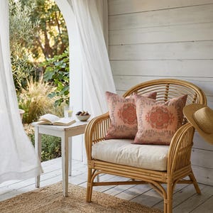 May include: A natural rattan armchair with two peach and rust patterned cushions. A straw hat hangs on the side. The chair is on a wooden deck, with a small table and a rug. A book and figs are on the table.
