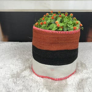 May include: A cylindrical woven basket featuring horizontal color-blocked stripes in rust orange, black, and off-white. The top and bottom edges are finished with a bright pink stitched trim. A small green plant with tiny orange flowers sits inside.