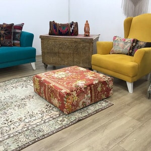 Pode incluir: Uma poltrona amarela com um almofada estampada com flores fica ao lado de um sof&aacute; azul turquesa com almofadas estampadas. Um pufe estampado com flores fica sobre um tapete estampado em creme e marrom na frente do sof&aacute;. Um ba&uacute; de madeira com entalhes intrincados fica atr&aacute;s do sof&aacute;.