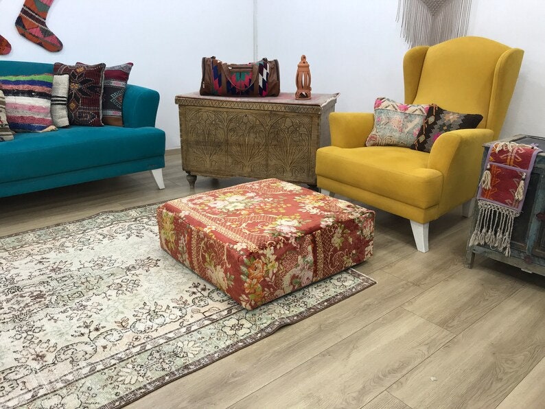 Pode incluir: Uma poltrona amarela com um almofada estampada com flores fica ao lado de um sof&aacute; azul turquesa com almofadas estampadas. Um pufe estampado com flores fica sobre um tapete estampado em creme e marrom na frente do sof&aacute;. Um ba&uacute; de madeira com entalhes intrincados fica atr&aacute;s do sof&aacute;.