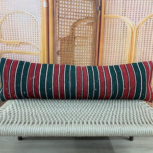 Puede incluir: Un cojín largo y rectangular con un estampado de rayas rojas, verdes y blancas. El cojín está colocado sobre un banco tejido de color beige claro. El fondo presenta una pantalla de madera.