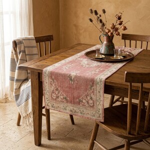 Puede incluir: Una mesa de comedor de madera está decorada con un camino de mesa con estampado floral en tonos rojos y crema. Una bandeja de metal contiene vasos y una jarra decorativa con flores secas. Una manta a rayas está sobre una silla de madera.
