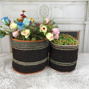 May include: Two cylindrical woven baskets in dark brown with horizontal cream stripes and orange stitched edges. The larger basket holds a bouquet of artificial blue, pink, and yellow flowers, while the smaller basket contains a green plant with small orange buds.