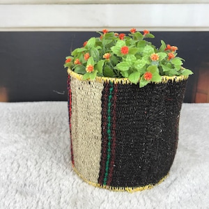 May include: A cylindrical woven basket featuring a striped pattern in black, beige, and thin green and red lines. The top and bottom edges are finished with yellow stitching. The basket contains a cluster of green foliage with small orange flowers.