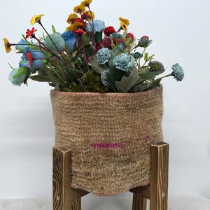 Könnte beinhalten: Ein brauner gewebter Korb mit einem rosa Streifen steht auf einem Holzständer. Der Korb ist mit einem Strauß künstlicher Blumen in den Farben Blau, Rot, Gelb und Grün gefüllt.