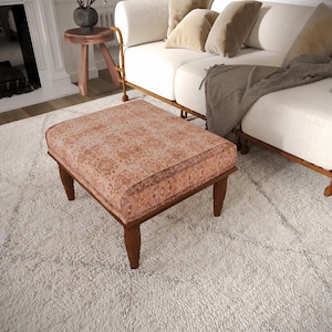 Pode incluir: Um otomano quadrado com um topo de tecido estampado em tons de terracota desbotado e creme. A peça é suportada por quatro pernas de madeira castanho-escura com um design cónico, proporcionando uma base estável para este móvel funcional.